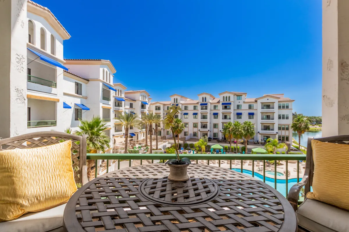 A peaceful balcony scene with cozy seating, overlooking the pool, palm trees, and neighboring buildings across the water. The inviting setup suggests a leisurely lifestyle—relaxing on the balcony, enjoying the pool, and connecting with the outdoors in a lush, sunlit environment.