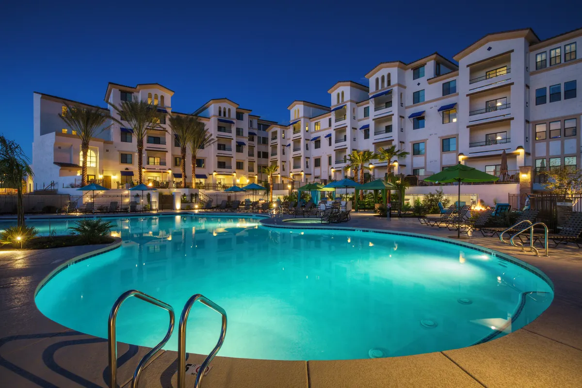 An inviting swimming pool is surrounded by palm trees, the building's facade, comfortable lounge chairs, and umbrellas. The scene suggests both leisure and social opportunities in a relaxed, visually appealing outdoor space.