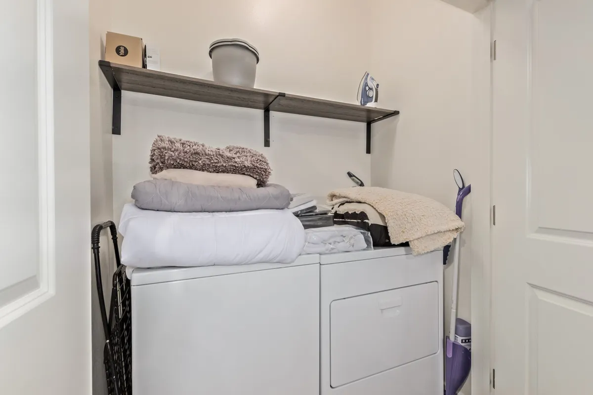 A practical laundry area with stacked clean linens, laundry appliances, and a shelf adorned with decorative items. The space is organized and homey, offering a glimpse of convenient in-unit amenities.