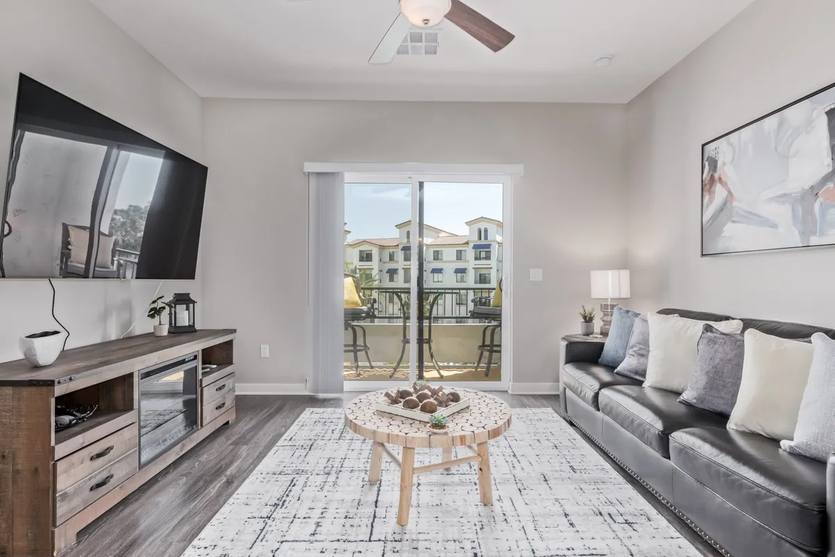 A stylish living room featuring a leather sofa, a modern coffee table with decorative items, a TV mounted on the wall, and a large sliding door that opens to a balcony, inviting in abundant natural light. The space looks ideal for both relaxation and entertaining.