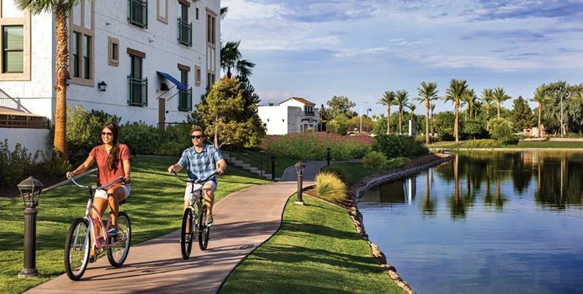 This exterior image shows bicyclists riding along a scenic lakeside pathway lined with palm trees, with a tranquil lake and resort-style building in the background. The scene is lively and active, emphasizing outdoor leisure and the proximity to nature.