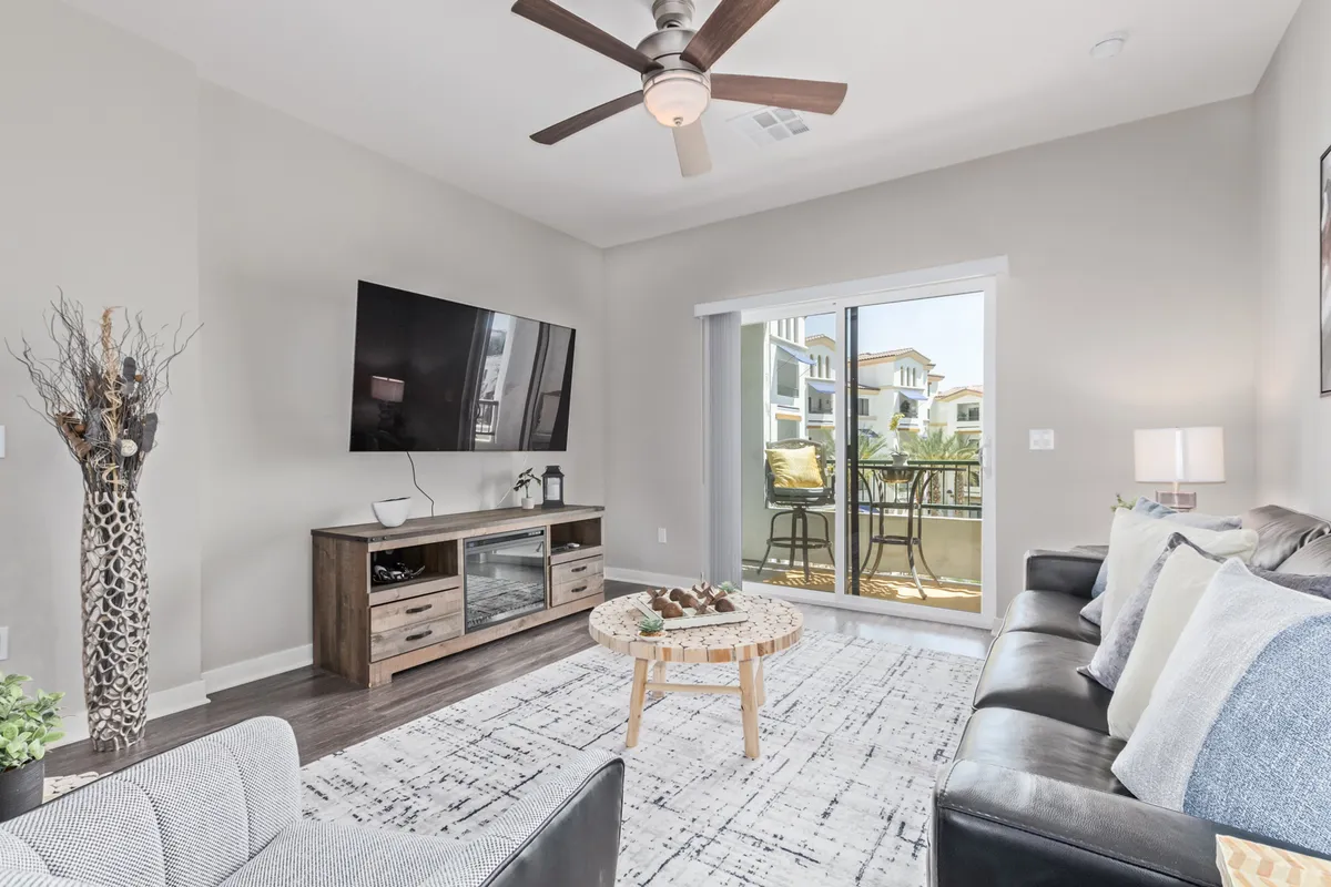 A cozy living room setup with a comfortable sofa, a coffee table, and a large TV. The space opens onto a balcony with views beyond, suggesting a connection to the outdoors and plenty of natural light. The mood is laid-back and stylish.