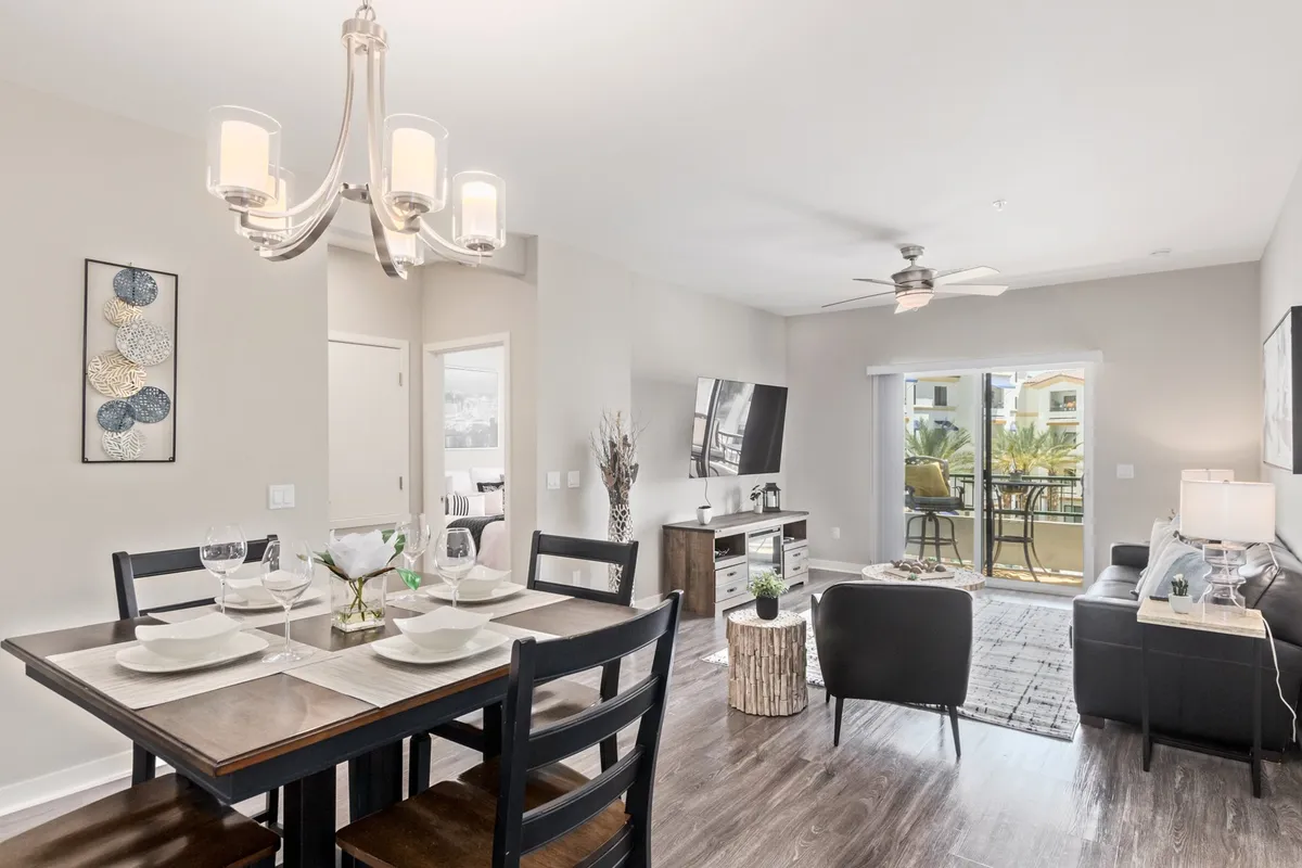 An inviting open-concept living and dining area that includes a dining table elegantly set for meals, a comfortable sofa area, a TV entertainment unit, and large balcony doors letting in natural light. The mood is warm and welcoming, perfect for gatherings.