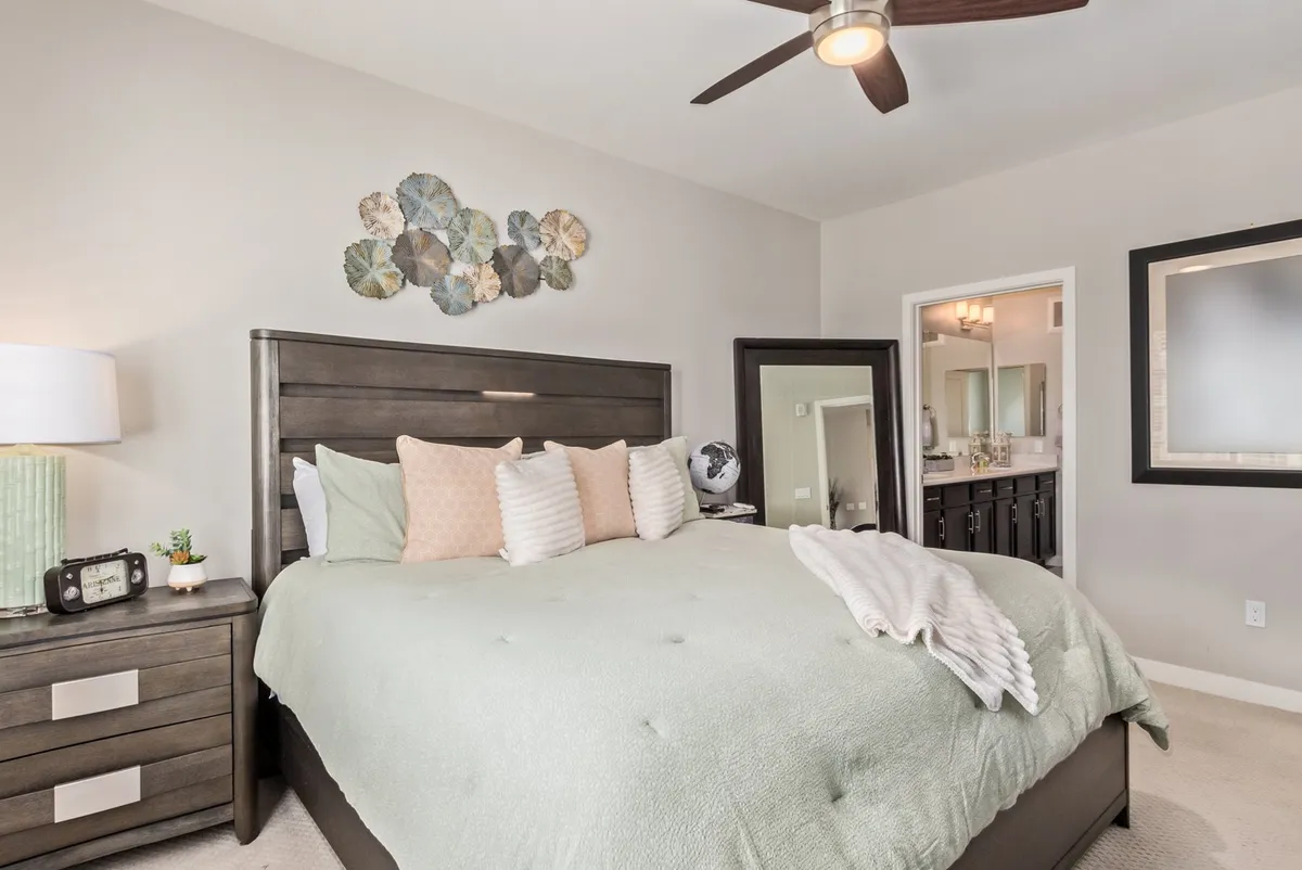 A cozy bedroom scene featuring a bed with soft green bedding, decorative wall art, and a nightstand with a lamp. Morning light gently filters in, creating a serene and inviting atmosphere ideal for rest and relaxation.