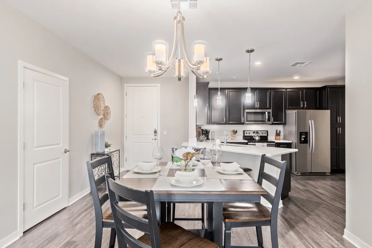 A warm, inviting dining area set for four, complete with a chandelier above and the kitchen visible in the background. Modern appliances and decorative accents create a cozy, intimate setting for meals and gatherings.