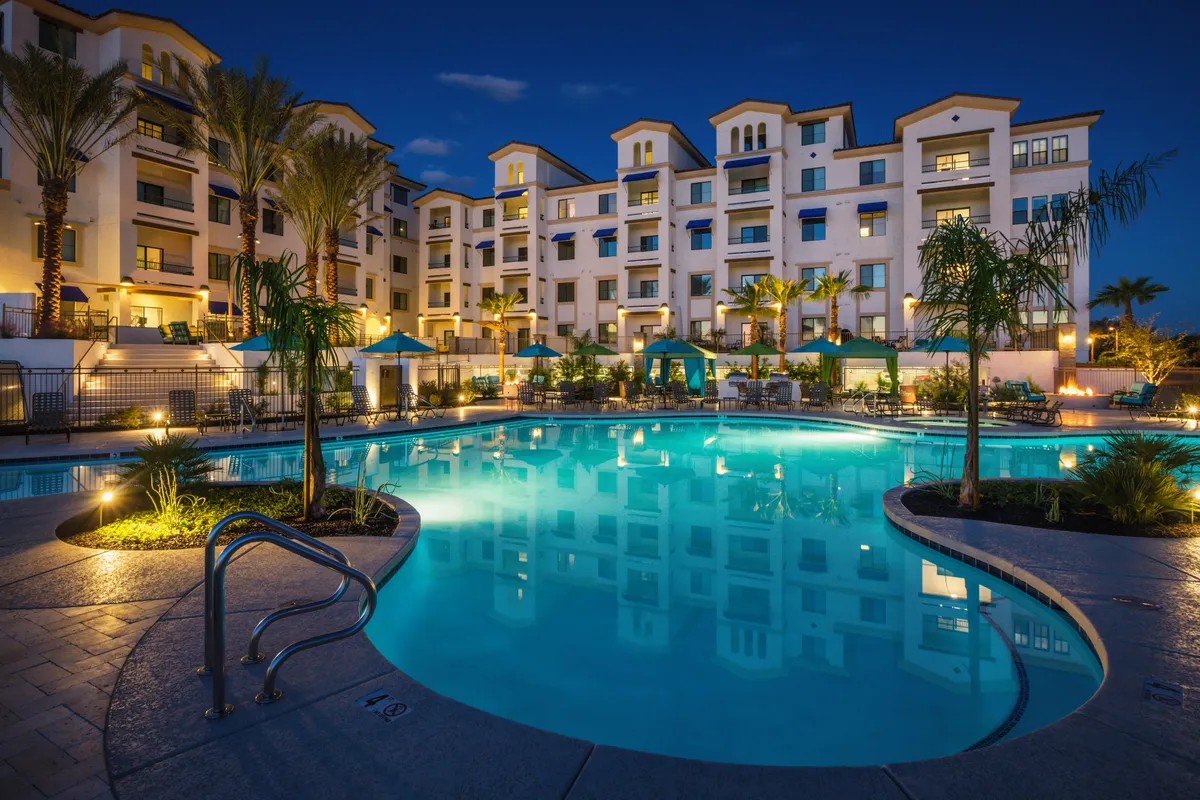 A sparkling swimming pool surrounded by elegant buildings, lush decorative palm trees, and cabana-style tents. Lounge chairs line the pool deck, inviting residents to relax, socialize, and soak up the Arizona sun. The tranquil water reflects a glowing sunset, and the setting exudes a luxurious, resort-like atmosphere.
