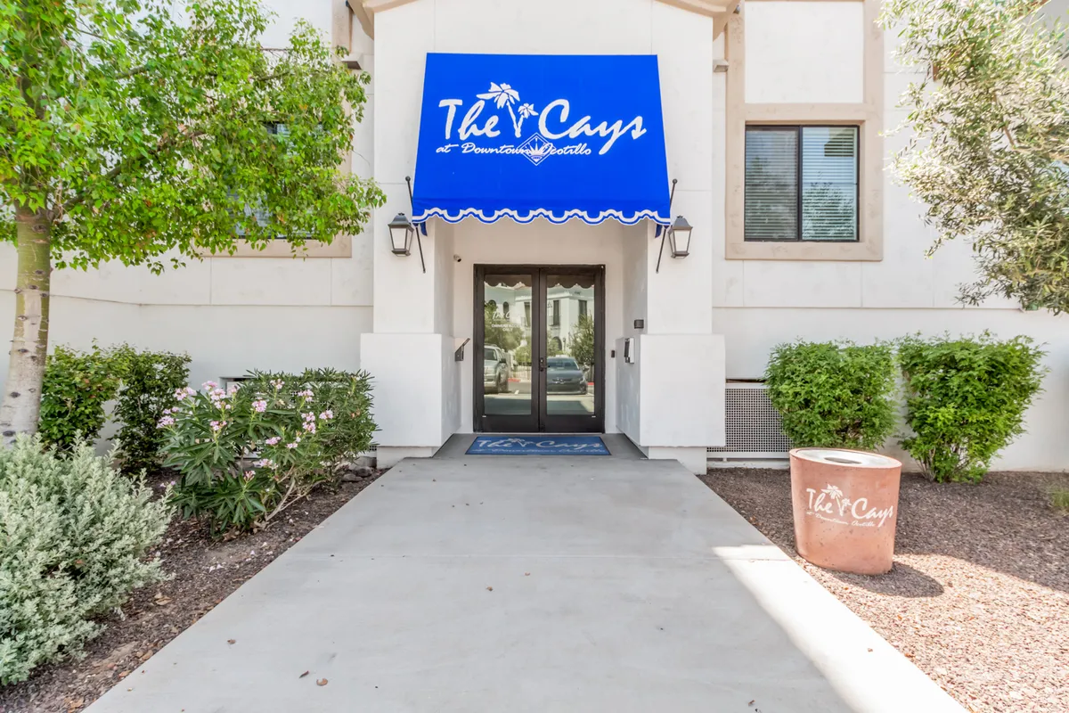 This exterior shot features the welcoming entrance with a blue awning branded 'The Cays', double glass doors, and manicured landscaping. The image conveys a friendly, resort-style atmosphere and sets the scene for a guest's arrival.