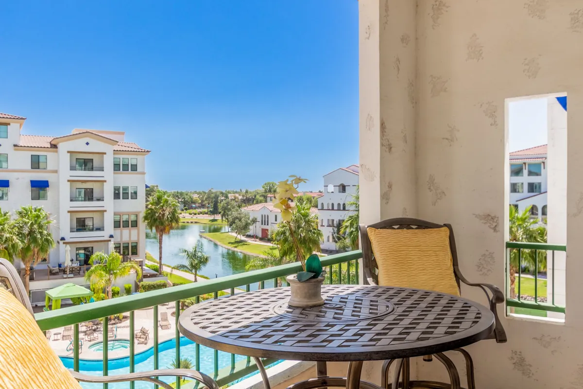 An exterior vantage from a balcony overlooking a serene pool area and waterway, framed by swaying palm trees. A table and chairs on the balcony suggest leisurely mornings or gathering with friends, while the view hints at tranquil canal-side paths and outdoor enjoyment.
